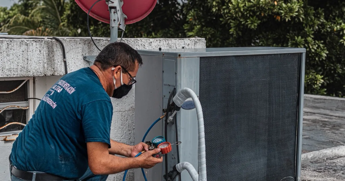A technician wearing a facemask connects manifold gauges to refill refrigerant on an outdoor AC condenser unit.