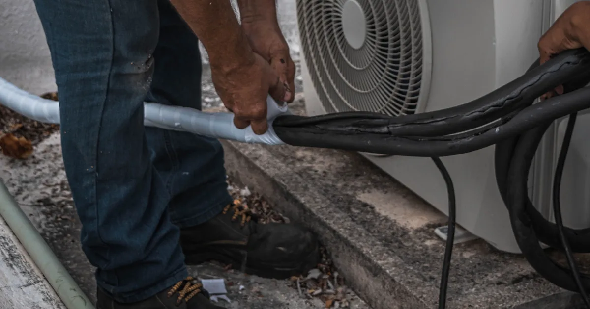 A technician tapes insulation around the pipe connections at the back of an outdoor air conditioning unit.