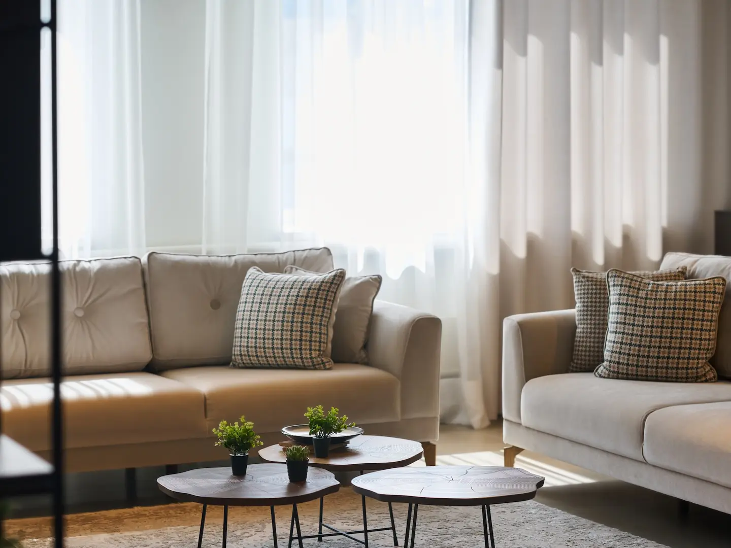 A sunlit living room with two cream sofas, houndstooth cushions and a cluster of wooden side tables in front of sheer curtains.