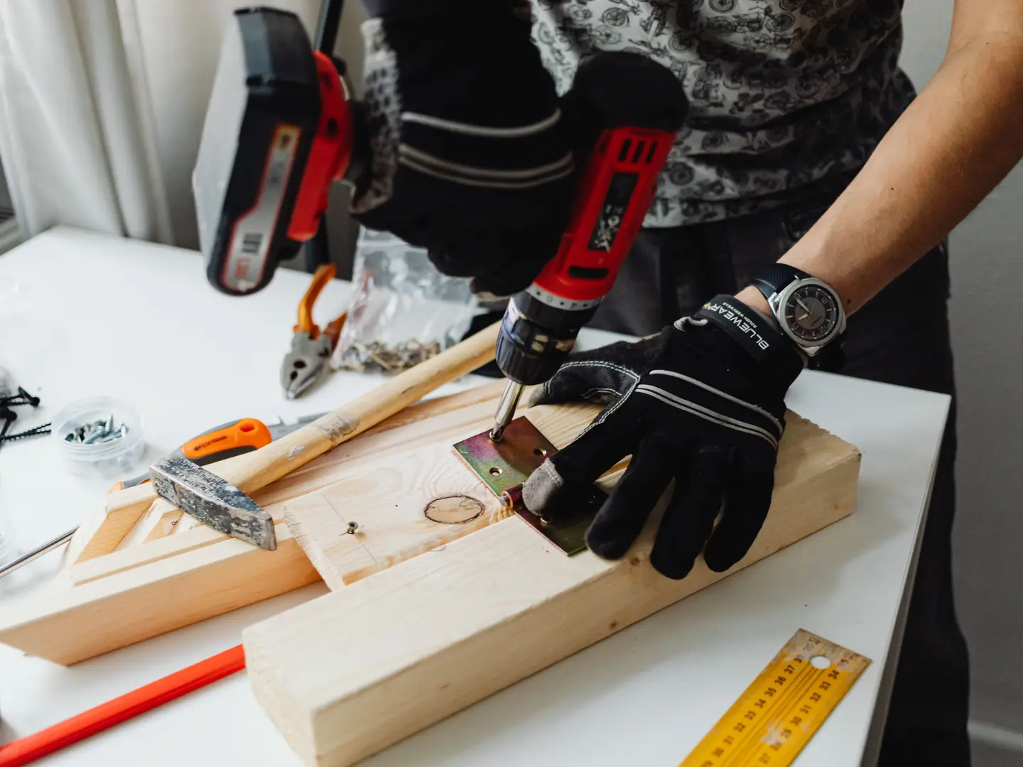 A gloved hand steadying a metal hinge on a length of pine while a cordless drill drives in the screw.